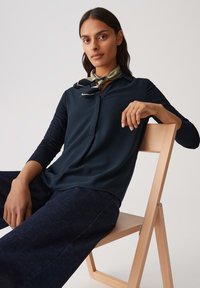 Navy blouse met een kraag en een halve knoopsluiting, gecombineerd met donkere denim. Model draagt een patroon sjaal knoop in de nek.
