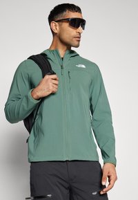 Chaqueta ligera con capucha en tejido verde de textura suave, con cremallera frontal y logo en el lado izquierdo del pecho, acompañada de una mochila negra.