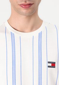 Fehér póló kék függőleges csíkokkal, kerek nyakkivágással és egy kis "Tommy Jeans" logóval a bal alsó részen. Pamut anyag.
