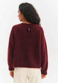 Burgundowy sweter z dzianiny o luźnym kroju, opuszczonych ramionach i fakturowanym wykończeniu. Na plecach znajduje się metka z napisem „Uważaj na słodkości”.