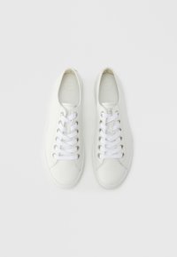 Baskets blanches avec une tige en cuir lisse, un design à lacets, des œillets métalliques, et une semelle en caoutchouc plate. Semelle intérieure texturée étiquetée "SUPER SOFT".