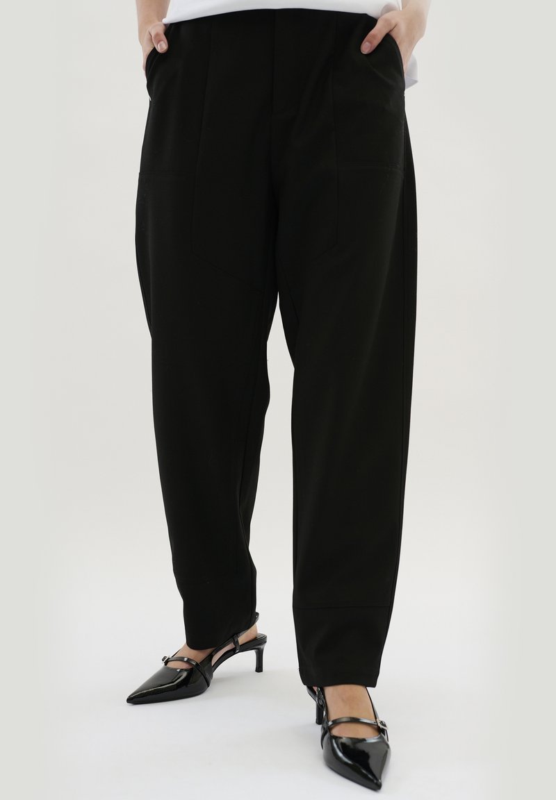 Personne portant un pantalon noir ample et des escarpins noirs à bout pointu avec bride arrière, debout les mains dans les poches devant un fond uni.