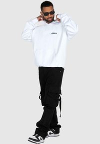 Hvid hoodie med en frontlomme og logo, parret med sorte cargo-bukser med flere lommer og stropper. Sorte og hvide sneakers.