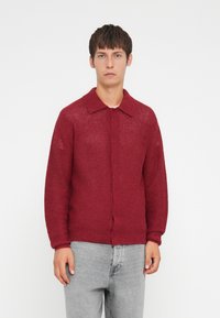 Red cardigan with a textured knit pattern, button closure, and a collar. Long sleeves and loose fit, paired with gray trousers.