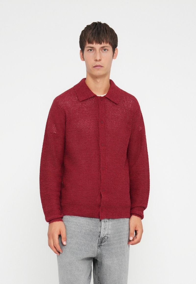 Red cardigan with a textured knit pattern, button closure, and a collar. Long sleeves and loose fit, paired with gray trousers.