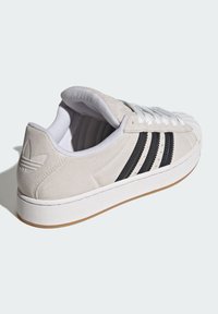 Svetlosivá semišová teniska s tromi čiernymi pruhmi, bielymi šnúrkami, bielym polstrovaným golierom a gumovou podrážkou. Na päte je vyrazené logo.