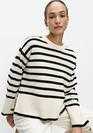 Kvinde med trukket hår tilbage iført guld øreringe og en hvid sweater med sorte vandrette striber, poserer foran en hvid baggrund.