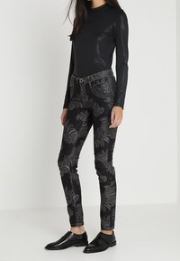 Černé přiléhavé kalhoty s květinovým vzorem stříbrnou nití. Hladká, elastická tkanina s vysokým pasem a zapínáním na knoflík. Černé kožené boty.