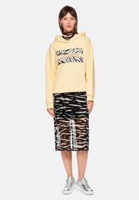 Gelber Kapuzenpullover mit schwarz-weißem Zebramuster, kombiniert mit einem transparenten schwarzen Rock mit ähnlichem Muster und silbernen Turnschuhen.