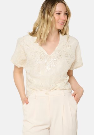 Beige sequined top with a V-neck and short sleeves, featuring a textured finish. Paired with light-colored tailored trousers.
