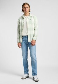 Hellgrünes kariertes Hemd über einem weißen T-Shirt, kombiniert mit blauen Jeans und zweifarbigen Sneakern. Das Hemd hat eine Brusttasche und eine Knopfleiste.
