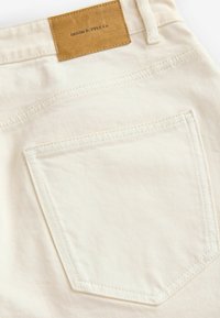 Hvide denim jeans med en baglomme i diamantform og et brunt lædermærke med teksten "DENIM SUPPLY Co." Synlige syninger.