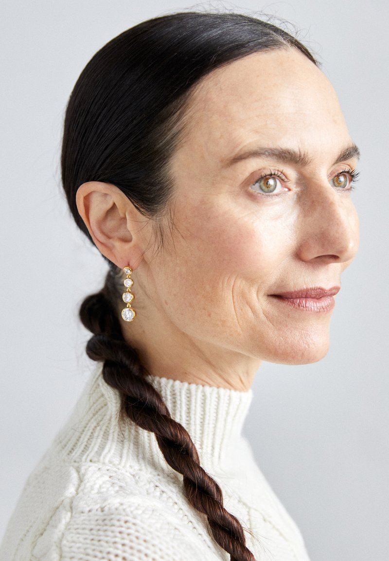 Gold earrings with three clear gemstones of varying sizes. The model has a braided hairstyle and wears a white, textured turtleneck sweater.