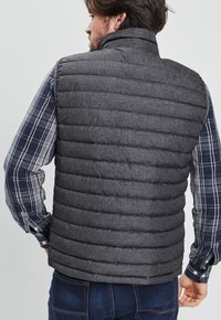 Chaleco gris acolchado con cuello alto, fabricado en un tejido texturizado y diseño de acolchado horizontal, llevado sobre una camisa de cuadros.