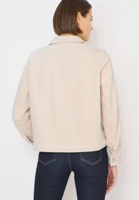 Beige kordfløjl jakke med krave, lange ærmer, søm over skulderbladet og manchetter med knapper. Bæres over mørke jeans med lyse syninger.
