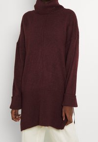 Maglione dolcevita bordeaux con finitura strutturata, polsini a coste e vestibilità comoda. Presenta spacchi laterali per facilitare i movimenti.