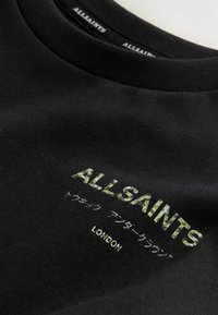 Juodas megztinis su tekstūruotu paviršiumi, kuriame yra žalias krašto stiliaus užrašas "ALLSAINTS" ir "LONDON" baltomis raidėmis. Apvalios apykaklės dizainas.