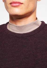 Burgundy texturerad tröja med ribbad halsringning, lager på lager över en beigefärgad polotröja. Närbild visar material och sömnadsdetaljer.