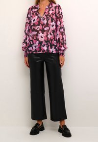 Kvinde iført en langærmet bluse i pink, lilla og sort med blomster, sorte bredde ben faux læderbukser og sorte loafers med gulddetaljer.