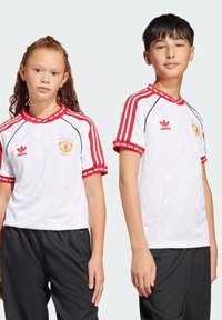 Bijele nogometne majice s crvenim detaljima, s kratkim rukavima, rebrastim ovratnikom i geometrijskom teksturom, uparene s crnim sportskim hlačama.