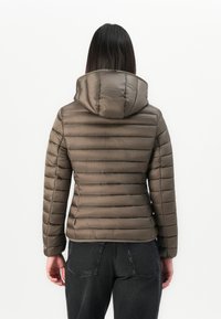 Vrouw met lang donker haar, gekleed in een bruine gewatteerde jas met capuchon, die met haar rug naar de camera staat tegen een eenvoudige witte achtergrond.