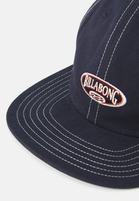 Marineblå cap laget av stoff med hvit søm. Har et brodert Billabong-logo i rødt og oval form på fronten.