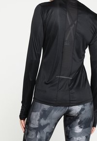 Černý sportovní top s dlouhým rukávem a síťovaným zadním panelem, doplněný šedými legínami s abstraktními vzory a čísly.