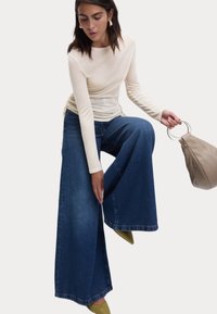 Beige langslavige top, blauwe wijde denim jeans, groenachtig gele schoenen, met een lichtbruine tas met een ronde handvat.