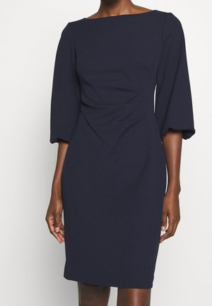 Femme portant une robe ajustée bleu marine foncé avec des manches trois-quarts et un décolleté bateau, debout devant un fond blanc uni.
