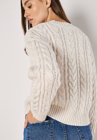 Cremfarbener Strickpullover mit Rundhalsausschnitt, geripptem Saum und strukturiertem Muster, kombiniert mit hochgeschnittenen blauen Denim-Jeans.