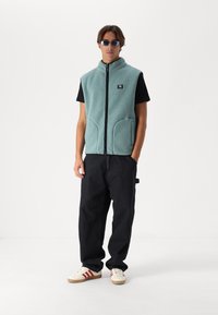 Vesta din fleece de culoare albastru deschis, cu guler înalt, închidere cu fermoar și două buzunare în față, asortată cu un tricou negru și pantaloni închisi la culoare.