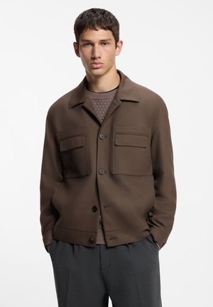 Ung mand iført en brun knapjakke med to forlommer med klap over en tekstureret sweater og mørkegrå bukser, hænder i lommerne.