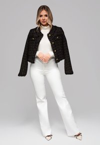 Frau trägt weißen Rollkragenpullover und Hose mit einer schwarzen, strukturierten kurzen Jacke und spitzen Pumps, steht vor einem grauen Hintergrund.