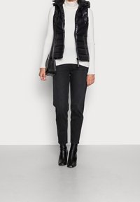 Zwarte gewatteerde bodywarmer met capuchon, gedragen over een witte geribbelde coltrui, gecombineerd met zwarte aansluitende jeans en enkellaarzen, en een kleine zwarte tas vasthoudend.