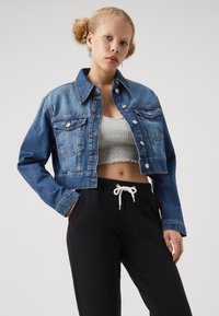 Cropped denim jacket in medium blue with two chest pockets, silver button closures, and a classic collar, paired with black sweatpants.