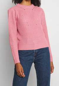 Pull en maille rose avec un motif texturé, doté d'un col rond, de manches longues et de petits détails découpés. Associé à un jean bleu foncé.