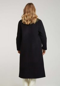 Manteau noir, long, avec une coupe droite et de longues manches, affichant un design simple et sans fermetures visibles. Le matériau semble doux.