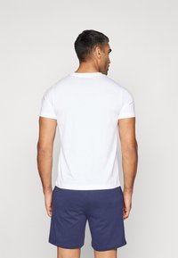 Camiseta blanca de algodón con mangas cortas, escote redondo y una silueta ajustada, combinada con pantalones cortos azul marino. Se muestra la vista trasera.