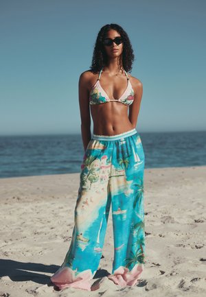 Femme portant un haut de bikini fleuri et un pantalon à jambes larges avec un imprimé plage et océan, debout sur une plage de sable sous un ciel dégagé.
