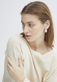 Boucles d'oreilles en argent en forme de goutte accrochées à un modèle aux cheveux longs, portant un pull léger beige avec des accents côtelés. Traits du visage subtils visibles.