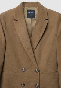 Blazer double boutonnage en velours côtelé marron avec un col à revers, deux grands boutons et une poche poitrine unique. Doublure intérieure beige.