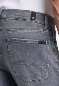 Vervaagde grijze denim jeans hebben een achterzak met een subtiel golvend stikpatroon, een zwarte merklabel en een knoopdetail op de zak.