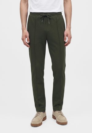 Homme portant un pantalon à cordon vert foncé et des chaussures beiges à lacets, debout devant un fond clair uni.
