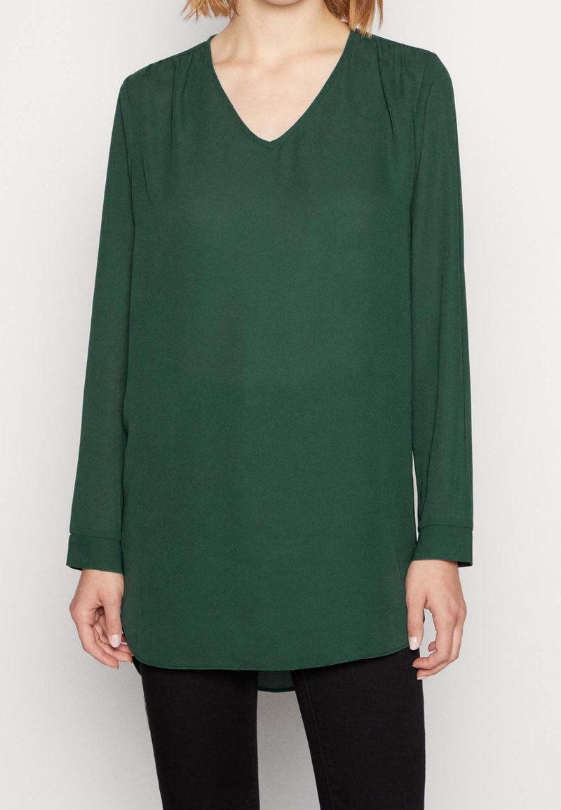 Långärmad blus med v-ringning i mörkgrön, tillverkad av slät tyg. Har subtila axelskärningar och en rak, något rundad nederkant.
