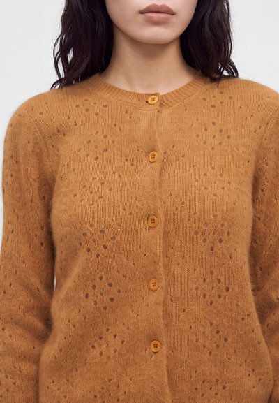 Femme portant un cardigan marron en maille duveteuse avec de petits trous en motif à pois et cinq boutons assortis, montré du cou à la taille.