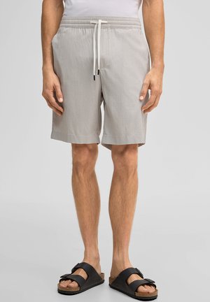 Man die lichtgrijze shorts met trekkoord en zwarte sandalen met dubbele band draagt, staand tegen een effen lichtgrijze achtergrond.