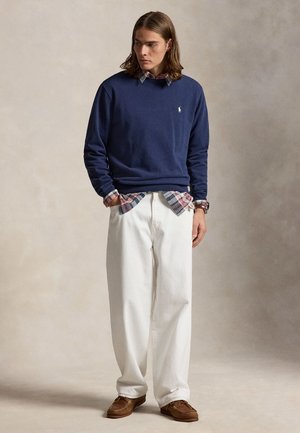Jeune homme debout, les mains dans les poches, portant un pull bleu marine sur une chemise à carreaux, un pantalon blanc à jambes larges et des mocassins marron.