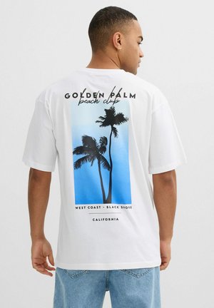 Hombre con camiseta blanca con gráfico de palmera azul y texto "Golden Palm Beach Club, Costa Oeste - Black Shore, California" en la espalda.