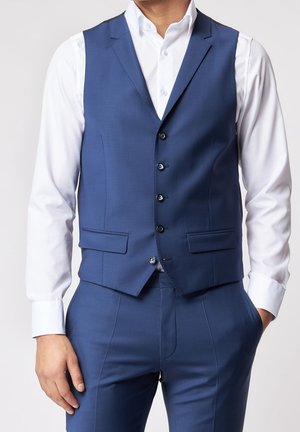 Man in een op maat gemaakt blauw pakvest en bijpassende broek met een wit overhemd, handen ontspannen langs het lichaam, tegen een effen achtergrond.
