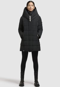 Cappotto piumino nero con grande cappuccio sovrapposto, design trapuntato, chiusura con zip, tasche laterali e logo accentato sul cappuccio.